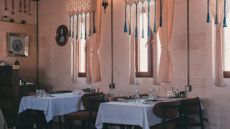 Beyaz duvarlı lokanta salonu, örtülü masalar ve pencere önünde dizili sandalyeler