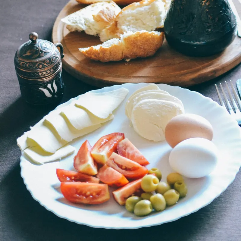 Beyaz tabakta peynir, domates ve zeytin; arkasında tahta tepside ekmek ve bakır cezve