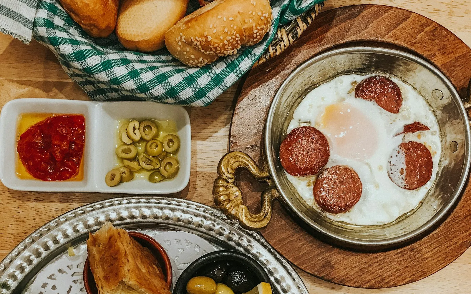 Ahşap masada bakır sahan içinde sucuklu yumurta, sepette simit ve ekmek, yanında zeytin ve acuka